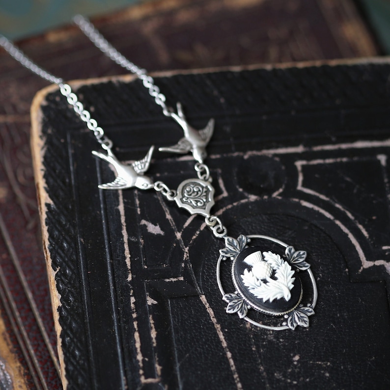 May include: A silver necklace with a black cameo featuring a white thistle. The necklace has two silver bird charms and a silver filigree charm.
