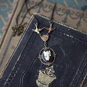 May include: A gold necklace with a black cameo pendant featuring a white angel. The necklace has two bird charms and a delicate chain.