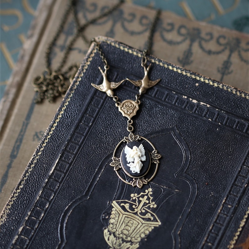 May include: A gold necklace with a black cameo pendant featuring a white angel. The necklace has two bird charms and a delicate chain.