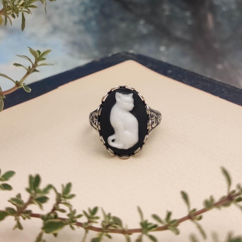 May include: A silver ring featuring an oval cameo with a white cat silhouette set against a black background. The metal band displays intricate, embossed scrollwork patterns along the sides, framing the central cat motif in a scalloped setting.