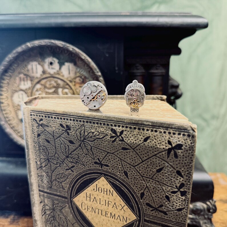 Puede incluir: Dos gemelos de estilo steampunk con engranajes de reloj expuestos. Uno es redondo, el otro tiene un dise&ntilde;o estilizado. Est&aacute;n sobre un libro antiguo con el t&iacute;tulo "John Halifax Gentleman". El fondo incluye una esfera de reloj y madera oscura.