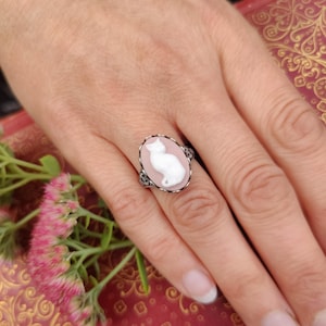 May include: An oval ring featuring a white cat cameo relief set against a soft pink background. The ring has a dark, textured metal band with intricate detailing around the edges of the oval setting, creating a distinct contrast.