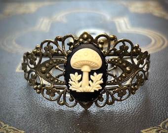 Puede incluir: Una pulsera antigua en tono latón con un camafeo ovalado que presenta un diseño de hongo color crema sobre un fondo negro. La pulsera tiene un diseño de filigrana ornamentado.