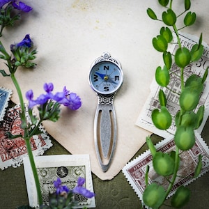 May include: A silver compass bookmark with a blue face and the words "JAPAN" and "W, E, S, N" printed on it. The bookmark is surrounded by vintage postage stamps and green leafy plants.