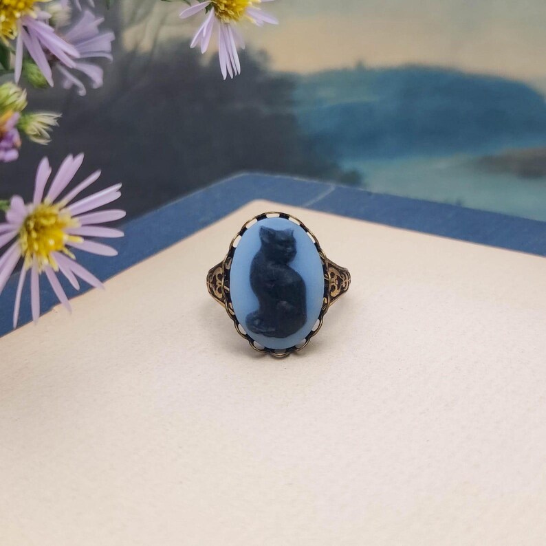 May include: An oval ring featuring a light blue cameo with a dark blue silhouette of a sitting cat. The cameo is set in an ornate, filigree-style antique gold-toned metal band with intricate scrollwork details along the sides of the setting.