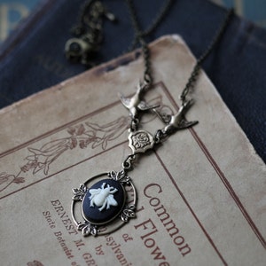 May include: A silver necklace with a cameo featuring a white bee on a black background. The necklace has two silver bird charms and a silver floral charm. The necklace is lying on a vintage book with the text "Common 1 Flower of sylvani by R. Gr."