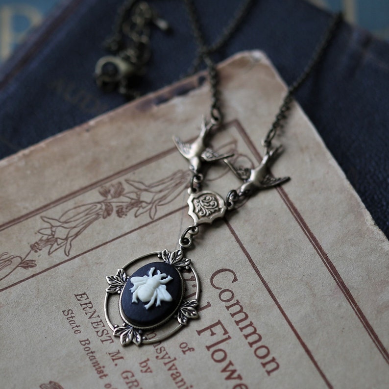 May include: A silver necklace with a cameo featuring a white bee on a black background. The necklace has two silver bird charms and a silver floral charm. The necklace is lying on a vintage book with the text "Common 1 Flower of sylvani by R. Gr."