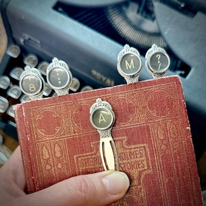 May include: A collection of silver-toned typewriter key bookmarks, each with a different letter or symbol, inserted into a vintage red book. The book has gold lettering that reads "Sherlock Holmes Stories". A vintage typewriter is in the background.