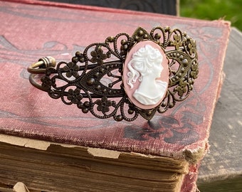 May include: A delicate bronze filigree bracelet with a cameo featuring a white silhouette of a woman's profile on a pink background.