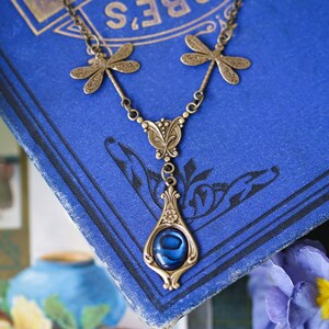 May include: A bronze necklace with a blue abalone shell pendant and dragonfly accents. The necklace is displayed on a blue book.