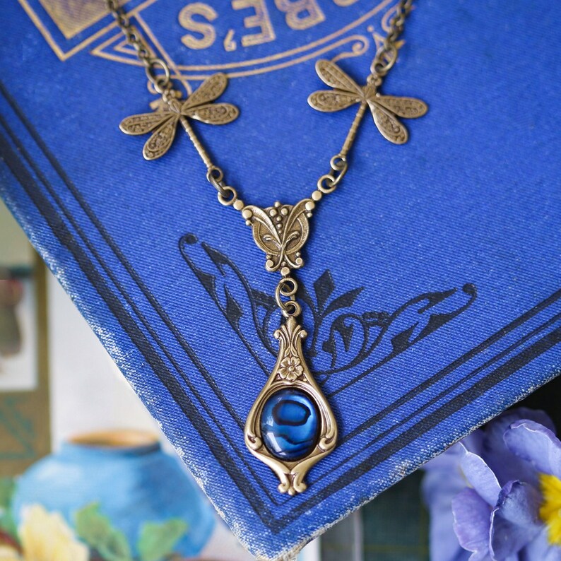 May include: A bronze necklace with a blue abalone shell pendant and dragonfly accents. The necklace is displayed on a blue book.