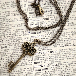 May include: A vintage-style brass key pendant necklace with a Celtic knot design. The necklace is hanging on a dictionary page with the word "murder" highlighted.