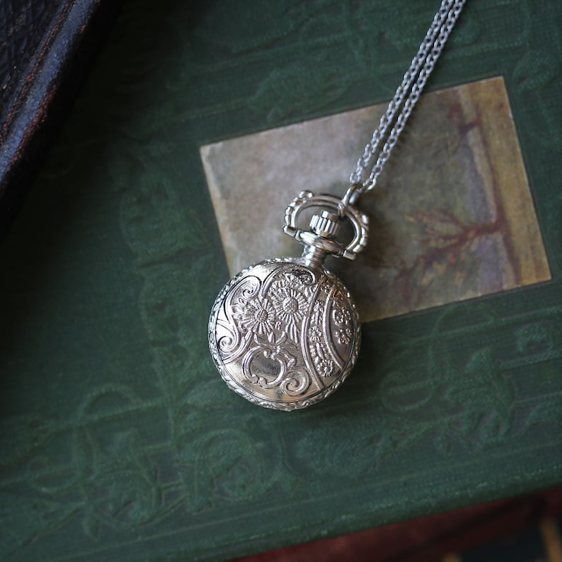 May include: A silver pocket watch with an ornate floral design hangs from a silver chain. The watch is on a green book with a gold-toned image.