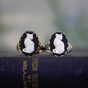 May include: Two oval rings featuring a white cat cameo set against a black background. One ring has a gold-toned metal band with ornate filigree detailing, while the other features a silver-toned metal band with similar intricate scrollwork.