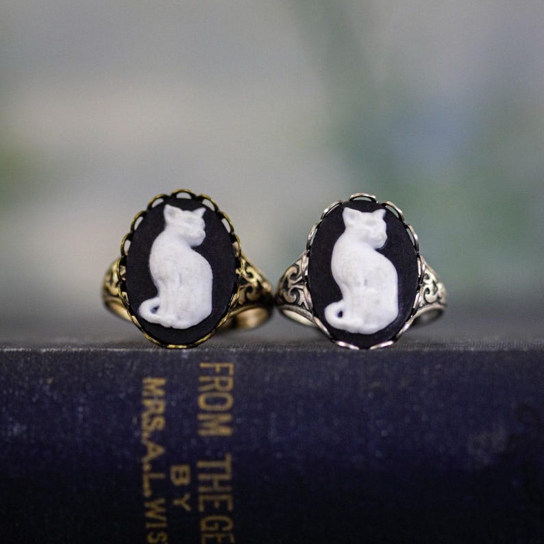 May include: Two oval rings featuring a white cat cameo set against a black background. One ring has a gold-toned metal band with ornate filigree detailing, while the other features a silver-toned metal band with similar intricate scrollwork.