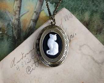 May include: A gold chain necklace with an oval locket featuring a white porcelain cat silhouette on a black background.