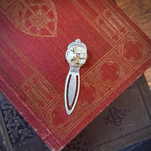 Puede incluir: Un marcap&aacute;ginas plateado con un dise&ntilde;o de mecanismo de reloj en la parte superior, con engranajes visibles y detalles intrincados. El marcap&aacute;ginas descansa sobre un libro rojo vintage con patrones dorados en relieve.