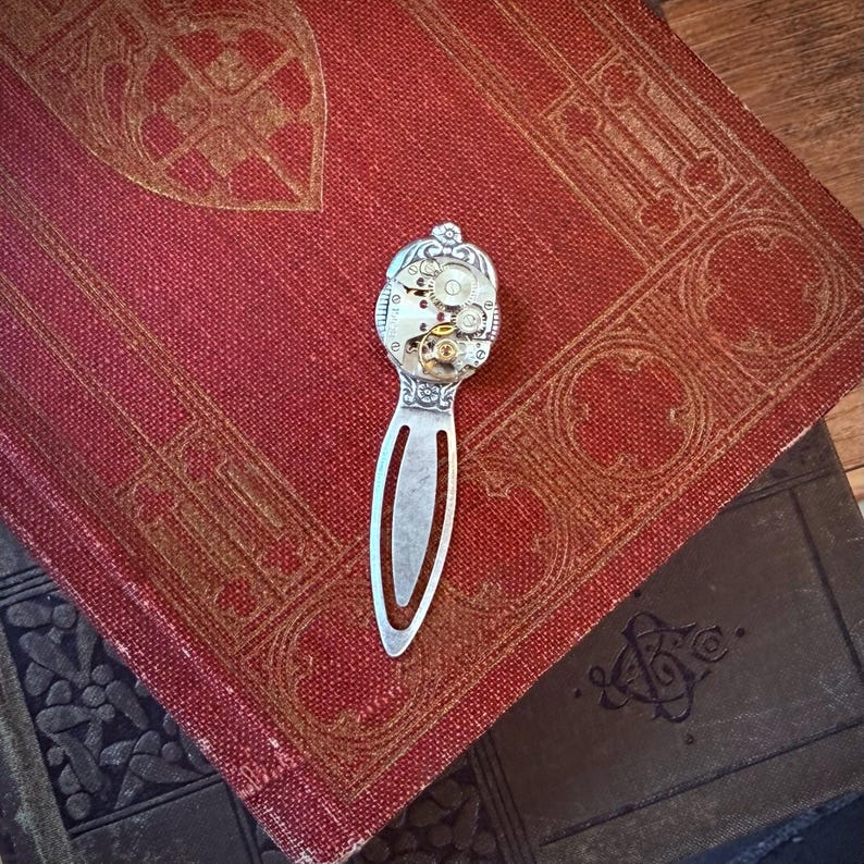 Puede incluir: Un marcap&aacute;ginas plateado con un dise&ntilde;o de mecanismo de reloj en la parte superior, con engranajes visibles y detalles intrincados. El marcap&aacute;ginas descansa sobre un libro rojo vintage con patrones dorados en relieve.