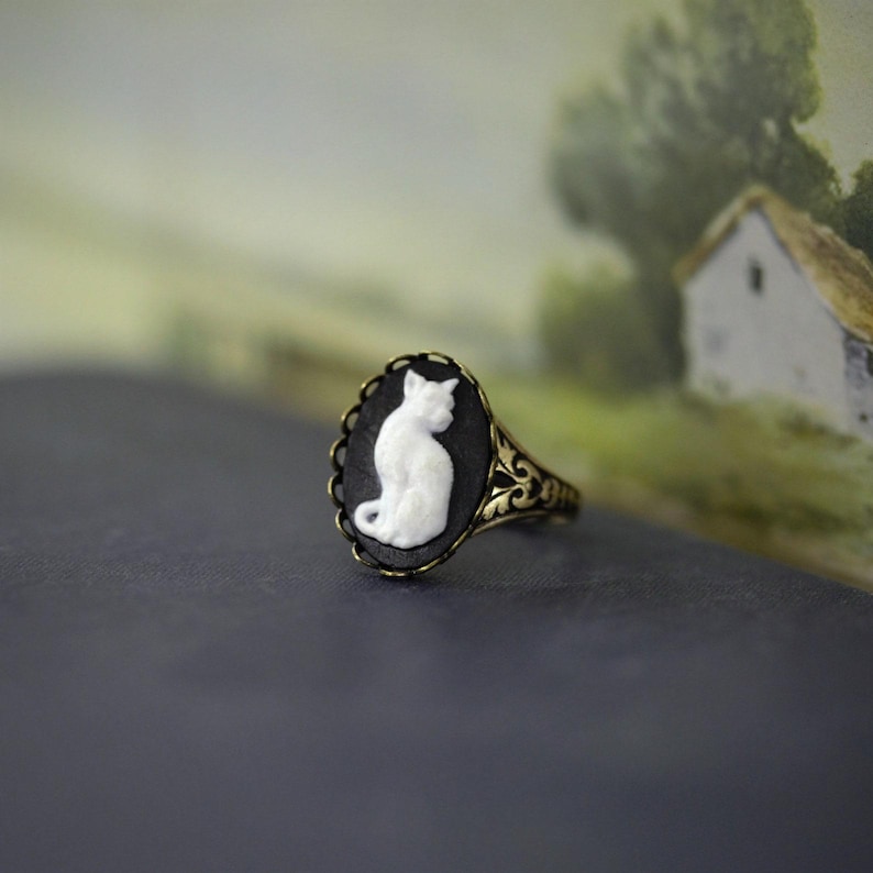 May include: An oval ring featuring a white cat cameo set against a black background. The ring has an antique brass-toned metal band with intricate filigree scrollwork detailing along the sides, framing the central cat motif.