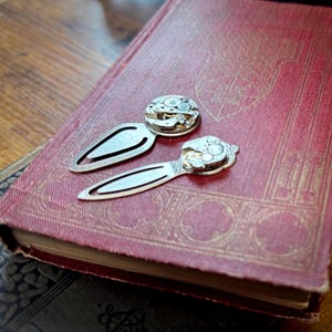 Puede incluir: Dos marcadores de metal plateado con detalles de movimiento de reloj sobre un libro rojo vintage. Los marcadores tienen un dise&ntilde;o cl&aacute;sico, con un mecanismo de reloj decorativo. El libro tiene patrones dorados en relieve.
