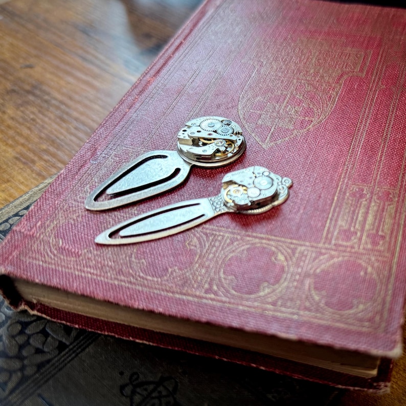 Puede incluir: Dos marcadores de metal plateado con detalles de movimiento de reloj sobre un libro rojo vintage. Los marcadores tienen un dise&ntilde;o cl&aacute;sico, con un mecanismo de reloj decorativo. El libro tiene patrones dorados en relieve.
