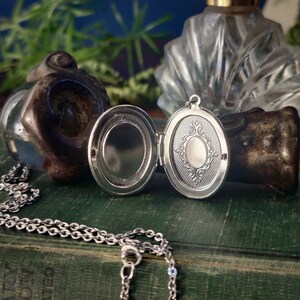 May include: Silver oval locket necklace with a detailed design on the front and a blank mirror inside. The locket is open and lying on a green book.
