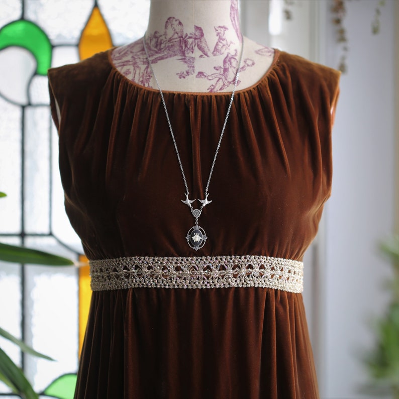 May include: A brown velvet dress with a lace trim at the waist. The dress features a silver necklace with a pendant that has two birds and a circular design.