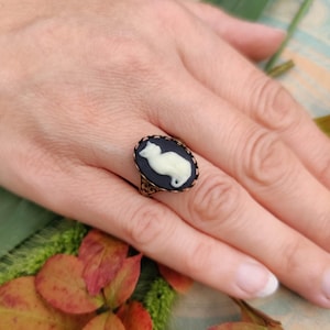 May include: An oval ring featuring a black cameo background with a raised white cat silhouette. The setting is made of a dark, filigree-style metal with intricate decorative patterns along the edges of the band and the oval frame.
