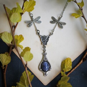 May include: A silver necklace with a blue gemstone pendant and dragonfly charms. The pendant is a teardrop shape with intricate details. The necklace is on a white background.