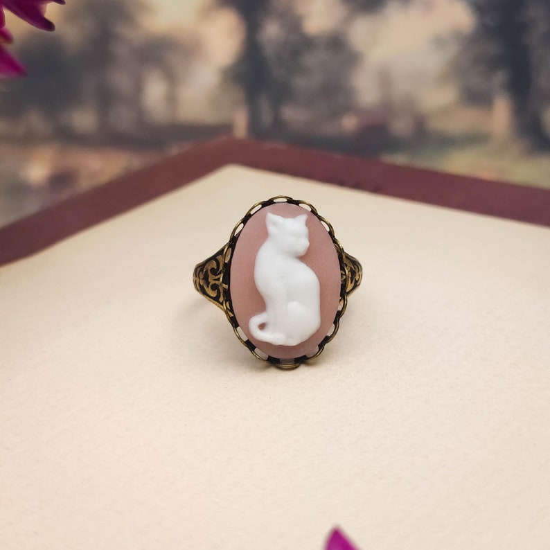 May include: An oval ring featuring a white cat cameo set against a soft pink background. The cameo is encased in an ornate, dark bronze-toned metal frame with intricate filigree detailing along the band and the edges of the setting.