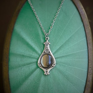 May include: A silver-toned pendant necklace with an oval-shaped, iridescent stone. The pendant has an ornate, floral design and hangs from a delicate chain. The necklace is displayed on a textured, light green surface.
