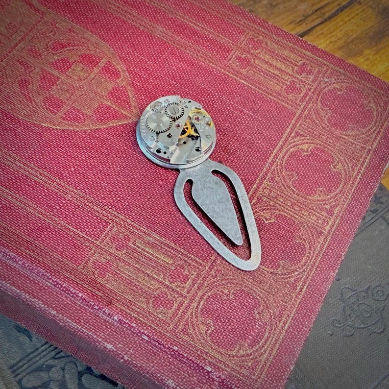 Puede incluir: Un marcap&aacute;ginas de metal plateado con un detalle de mecanismo de reloj. El marcap&aacute;ginas est&aacute; sobre un libro rojo con dise&ntilde;os dorados en relieve. El mecanismo del reloj presenta peque&ntilde;os engranajes.