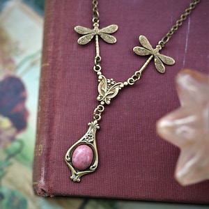 May include: A gold necklace with a pink stone pendant and dragonfly charms. The necklace is on a burgundy background.
