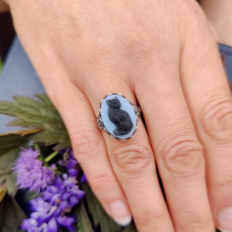 May include: An oval ring featuring a black cat cameo set against a light blue background. The cameo is framed by a silver-toned metal setting with a scalloped edge, creating a distinct contrast between the dark feline figure and the pale base.