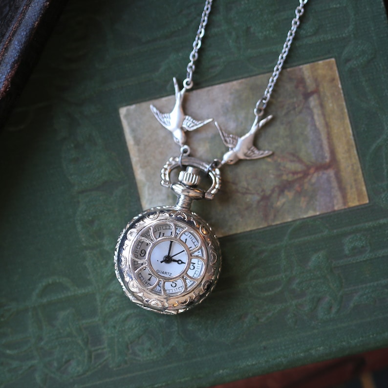 May include: Silver necklace with two silver bird charms and a silver pocket watch with a white face and black hands. The watch face has Roman numerals and the word "QUARTZ" printed on it.