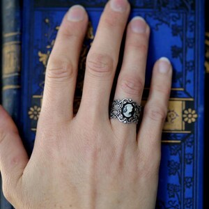 Pode incluir: Um anel de prata com um camafeu preto e branco com o perfil de uma mulher. O anel est&aacute; em uma m&atilde;o contra um fundo azul.