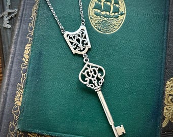Könnte beinhalten: Eine silberne Halskette mit einem Schlüsselanhänger und einem dekorativen filigranen Anhänger. Die Halskette wird auf einem grünen Buch mit goldenem Design gezeigt.