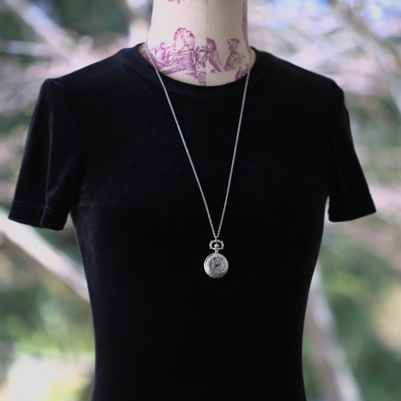 May include: A silver pocket watch necklace with a white face and black hands. The watch is on a silver chain and is hanging from a black velvet mannequin.