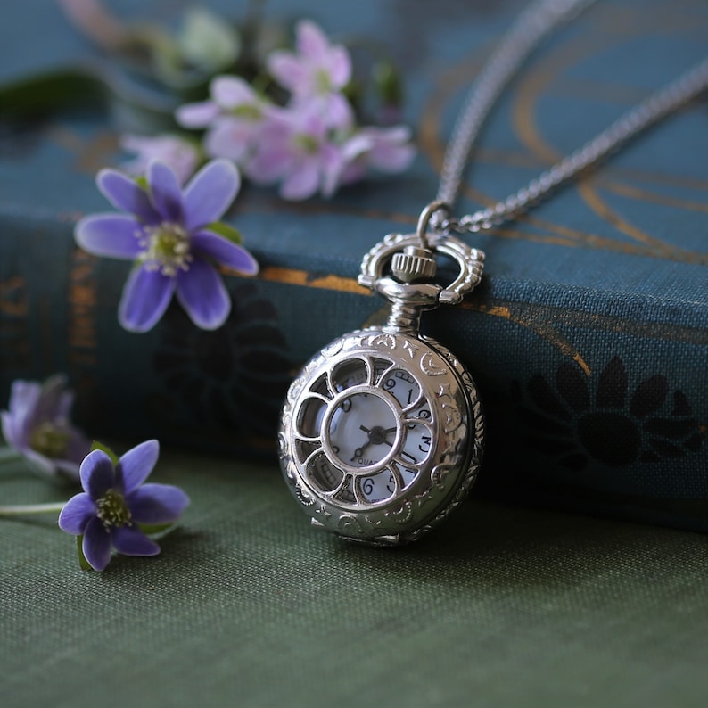 May include: A silver pocket watch necklace with a delicate chain and an ornate design. The watch face has a white background with black Roman numeral markings and a silver hand.