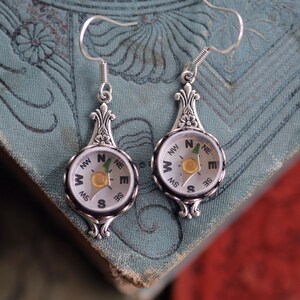 Puede incluir: Pendientes de plata en forma de brújula con borde negro y cara blanca. La cara de la brújula tiene las letras N, S, E, W, NW, NE, SW, SE y una pequeña flecha verde que apunta al norte.