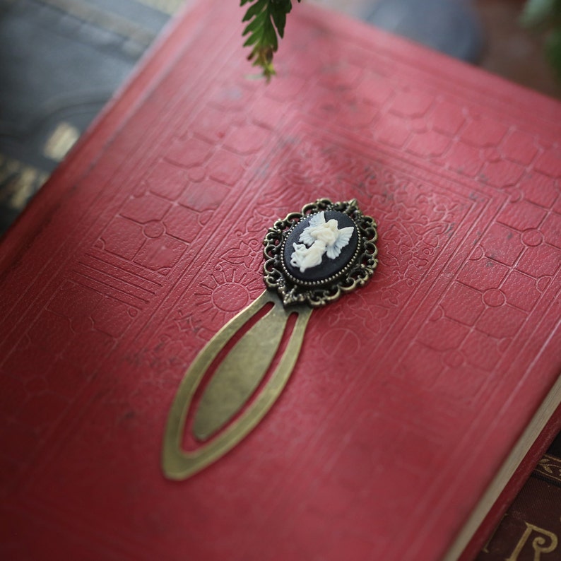 May include: A bronze bookmark with an ornate design features a cameo of a white angel with black wings. The bookmark is on a red book cover.