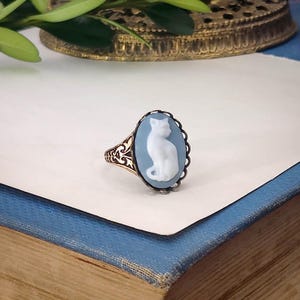 May include: An oval ring featuring a light blue cameo with a white cat silhouette in profile. The setting is made of an intricate, dark metallic filigree band with a scalloped edge surrounding the central cameo stone.