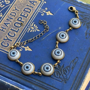 May include: A gold chain bracelet with six blue glass eyes. The eyes have white scleras and black pupils. The bracelet is on a blue book with gold lettering.