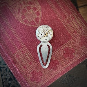 Puede incluir: Un marcap&aacute;ginas de estilo steampunk con un dise&ntilde;o plateado alargado y una parte superior circular. La parte superior est&aacute; adornada con intrincados engranajes y mecanismos de reloj. El marcap&aacute;ginas descansa sobre un libro vintage de color burdeos con patrones en relieve.