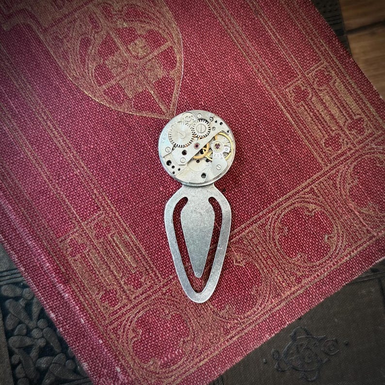 Puede incluir: Un marcap&aacute;ginas de estilo steampunk con un dise&ntilde;o plateado alargado y una parte superior circular. La parte superior est&aacute; adornada con intrincados engranajes y mecanismos de reloj. El marcap&aacute;ginas descansa sobre un libro vintage de color burdeos con patrones en relieve.