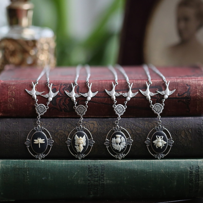 May include: Five silver necklaces with black cameo pendants featuring a dragonfly, angel, thistle, and bee. Each necklace has a pair of silver birds flying above the pendant.