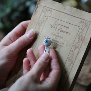 May include: A silver bookmark with a blue glass eye charm. The bookmark is being held by a hand and is resting on a book titled "Common Wild Flowers of Pennsylvania".