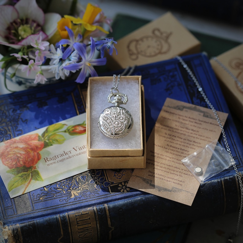 May include: A silver pocket watch with a floral design on the face, hanging from a silver chain. The watch is in a small brown box, and there is a small card with instructions on how to set the watch.