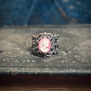 Pode incluir: Um anel de prata com um design ornamentado que apresenta um camafeu com o perfil de uma mulher em rosa.
