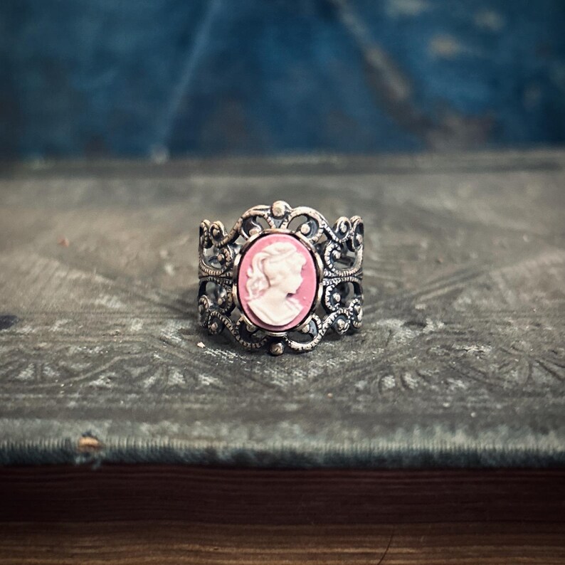 Pode incluir: Um anel de prata com um design ornamentado que apresenta um camafeu com o perfil de uma mulher em rosa.